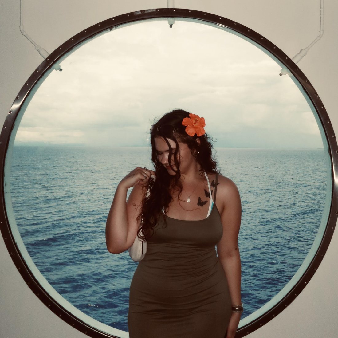 Hannah in a round ship window over the open ocean, a hibiscus in her hair